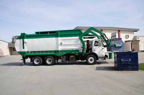 A large commercial waste collection truck from Commercial Waste Camden parked on a paved driveway in Camden, with a green and white colour scheme and a hydraulic lifting arm positioned above an open blue large-capacity skip bin filled with waste. The truck has a sturdy design with a metallic finish, surrounded by a modern residential building with white walls and red window frames in the background. The environment appears clean and organized, with the truck ready for rubbish removal services in the local area, reflecting a professional approach to waste disposal for commercial or large-scale residential clients. The scene is well-lit with natural daylight, emphasizing the functional and industrial appearance of the waste removal vehicle and the adjacent waste bin, aligning with the company's service offerings for rubbish collection in the Camden region.