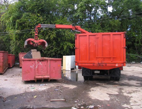 A red hydraulic waste compactor attachment with a claw mechanism is shown lifting debris into a large, red, metal waste container positioned on a paved surface. The attachment is connected to an arm extending from a vehicle that is partially visible on the right side of the image. The background features green trees and foliage, indicating an outdoor setting. The waste container is situated on an asphalt or concrete ground with some scattered litter and small debris nearby, suggesting a site for rubbish removal or waste management services. The scene appears to be part of a commercial waste removal operation, with the equipment and containers used by a company such as Commercial Waste Camden operating within Camden or a similar area. The lighting is natural, with daylight illuminating the scene clearly.
