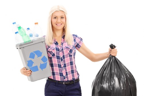 A young woman with long blonde hair and a friendly expression, dressed in a short-sleeved, pink and blue plaid shirt and dark trousers, stands against a plain white background. She holds a grey recycling bin in her left hand, which is filled with empty plastic bottles of various colors, including green, clear, and blue, and some with orange caps. In her right hand, she grips a large black rubbish bag made of thick plastic material, appearing full and tied at the top. The composition suggests a scene related to waste disposal or rubbish collection, emphasizing recycling and rubbish management. The image is bright, well-lit, and captures the thematic focus on waste removal services, subtly resonating with local rubbish collection practices in areas like Camden, aligning with the services offered by Commercial Waste Camden.