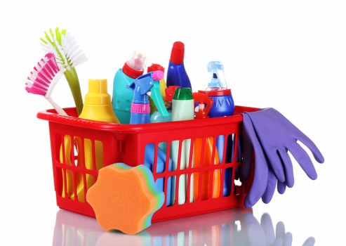 A red plastic basket filled with various cleaning supplies and equipment, including spray bottles, scrub brushes, rubber gloves, and a sponge, all arranged on a flat surface. The spray bottles are in multiple colours such as blue, red, and green, with some featuring trigger nozzles. The scrub brushes have brightly coloured bristles, with one in pink and another in green. The rubber gloves are purple, folded over the edge of the basket, positioned on the right side. In front of the basket is an orange cleaning sponge with a rainbow outline, resting on the surface. The background is plain white, emphasizing the colourful assortment of cleaning tools. This image visualises supplies commonly used in professional or domestic rubbish and waste removal and cleaning services, aligning with the context of commercial waste management in Camden, UK, provided by Commercial Waste Camden.