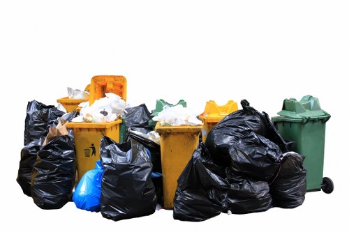A collection of multiple rubbish waste bins and large black garbage bags arranged outdoors, likely on a driveway or pavement area. The waste bins are made of heavy-duty plastic with textured surfaces and are in various colours including yellow, green, and grey, with some lids open or partially open. Surrounding the bins are numerous black plastic bags filled with waste, some tied at the top, with visible crumpled textures. A bright blue bag is also among the waste, contrasting with the darker bags. The background is plain white, indicating a studio or digitally isolated image, with no visible environmental context or specific location details. The arrangement demonstrates typical waste collection materials used by commercial waste management services such as Commercial Waste Camden, contributing to the regular rubbish removal activities in Camden or nearby areas.