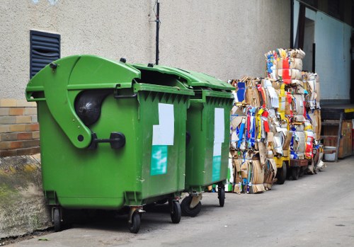 A green underground refuse bin with a closed lid, situated on a paved pathway alongside a concrete building wall in Camden. The bin is made of durable plastic with a textured surface and has white rectangular labels attached to its front. To the right of the bin, there are large compacted bundles of cardboard and paper packaging stacked against the building, showing various colours from printed cardboard boxes and food packaging. The area appears to be an urban back alley or service access point, with the ground being smooth and slightly worn. The background includes a vent on the wall and a partially visible glass window or door frame. The scene illustrates typical waste collection and disposal management practices relevant to commercial properties, with arrangements handled by companies like Commercial Waste Camden in the Camden post code area, contributing to effective rubbish management strategies in the town.
